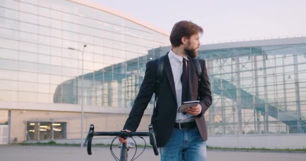 Guapo confiado joven exitoso gerente de la oficina barbuda va con su bicicleta cerca de edificio de vidrio urbano y el uso de i-pad, cámara lenta — Vídeos de Stock