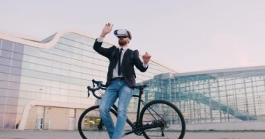 Kendine güveni tam, 30 yaşında, sanal gerçeklik kulaklığı takmış bir iş adamı bisikletinin yanında duruyor ve modern şehir binasının arka planında hayali bir ekran üzerinde çalışıyor.
