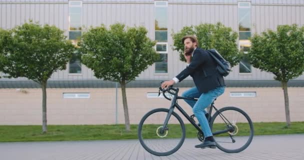 Πλευρική άποψη του όμορφος χαμογελαστός νεαρός γενειοφόρος άνδρας με κομψά ρούχα που ιππασία ένα ποδήλατο και έχει θετική τηλεφωνική συνομιλία — Αρχείο Βίντεο