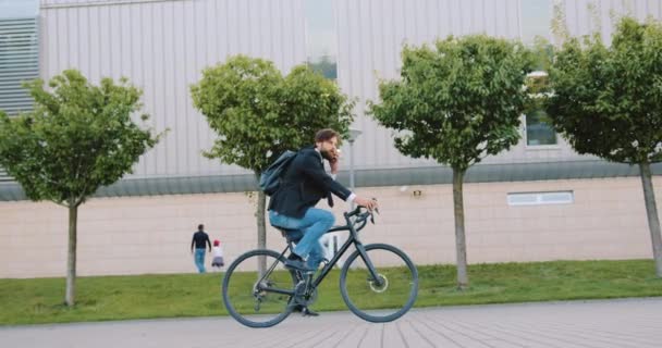 都市部の近代的な建物の背景に自転車に彼の仕事に到達しながら、電話で話す魅力的な自信を持って若いひげのあるオフィスの労働者 — ストック動画