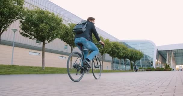 Good-looking stylish contented young bearded guy riding a bicycle to his work on cobbled path near modern big bulding with glass windows,slow motion — Stock Video