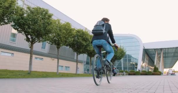 Ймовірно, концентрований молодий бородатий офісний працівник з рюкзаком їде на велосипеді на забитій вулиці біля сучасного будівництва аеропорту з панорамними вікнами, повільний рух — стокове відео