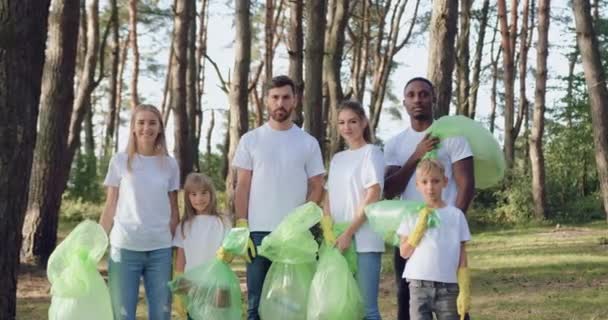 Atractivo grupo multiétnico de jóvenes y niños posando en la cámara después de limpiar el parque de la dispersión de basura en bolsas de plástico, de cerca — Vídeos de Stock