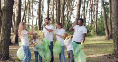 Çekici gülümseme portresi. Yetişkinler ve çocuklardan oluşan karışık bir yarış takımı. Parktaki tüm plastik torbalara çöp topladıktan sonra beşlik çakıyorlar.