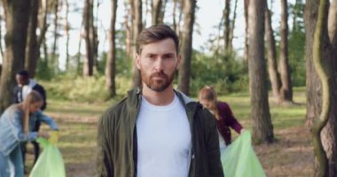 Parkta çöp toplayan gönüllü arkadaşlarının arka planında kameraya poz veren konsantre sakallı adamın portresi.