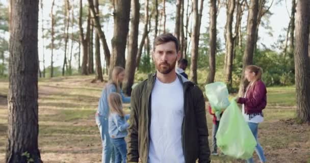 Good-looking smiling bearded man standing in the background of park where his friends-activists collecting rubbish into plastic packs all over the park — Stock Video