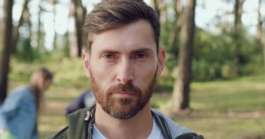 Çekici, yoğun sakallı bir adamın ön görüntüsü. Arka planda, çöpleri parkta plastik poşetlere toplayan bir grup insanın görüntüsü.