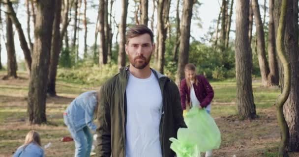 Retrato de un hombre barbudo serio y guapo posando en cámara con una bolsa de basura que recogió junto con otra gente por todo el parque — Vídeo de stock