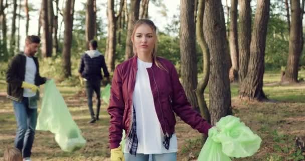 Primer plano de chica activa seria y guapa que de pie frente a la cámara con la bolsa de basura durante la recolección de todo el parque junto con voluntarios de ideas afines — Vídeos de Stock