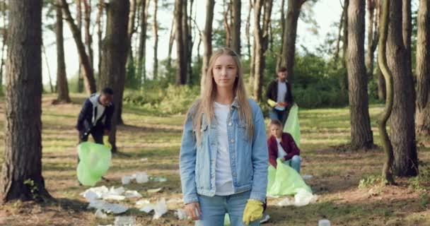 Belle fille confiante souriante debout devant la caméra sur le fond du groupe de personnes-bénévoles qui collectent les ordures dans le parc, concept de protection de l'environnement — Video