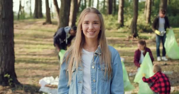 Adorable satisfied smiling young woman posing on camera near volunteers from eco team which with plastic bags collecting trash in the park,society against pollution — Stock Video