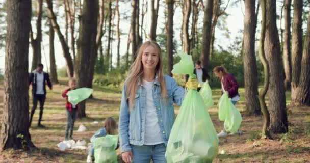 Bastante sonriente chica trabajadora con bolsa de basura con diferentes residuos posando en la cámara cerca de sus amigos-como-mentalidad que recoge la basura en todo el parque — Vídeos de Stock