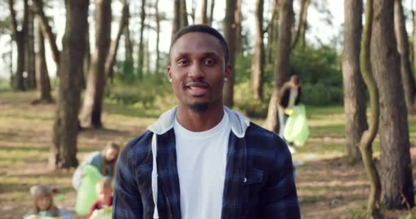 Portrait of good-looking smiling satisfied black-skinned male activist which looking at camera with thumbs up while cleaning park territory from rubbish together with friends-like-minded — Stock Video