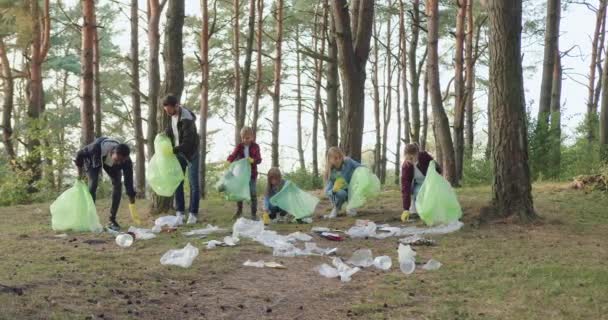 Belle équipe positive active jeune multiraciale active d'adultes et d'enfants dans des gants de protection ramassant les déchets dans le parc dans des sacs en plastique, concept de société contre la pollution — Video