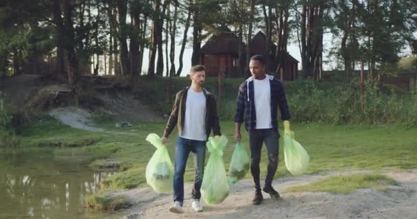 Красиві задоволені змішані гонки молоді чоловіки носять поліетиленові пакети біля озера після очищення навколишньої території від сміття, вид спереду — стокове відео