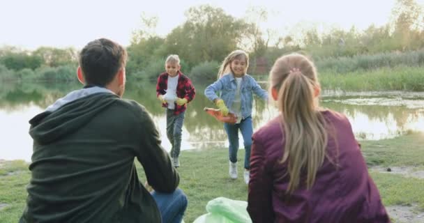 Dobrze wyglądający zadowoleni młodzi rodzice siedzący razem na plaży nad jeziorem i obserwujący jak ich dzieci zbierają śmieci i wkładają je do plastikowych toreb. — Wideo stockowe