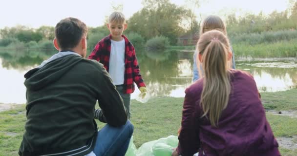 Likable uśmiechnięty szczęśliwy chłopiec i dziewczyna pomagając rodzicom zbierać śmieci z brzegu jeziora i dając przybić sobie piątkę — Wideo stockowe