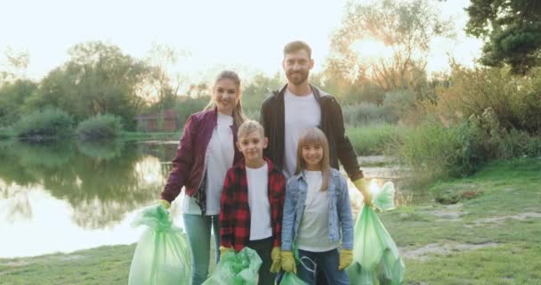 Vista frontal de una atractiva familia joven y sonriente con niños que se paran frente a la cámara con bolsas de basura después de limpiar el territorio de los lagos — Vídeos de Stock