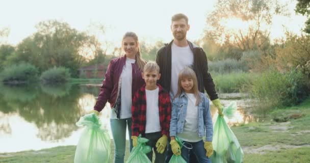 Adorable familia alegre feliz de los padres y los amantes de la naturaleza de los niños posando en la cámara con bolsas de basura de plástico después de limpiar el territorio circundante cerca del lago — Vídeos de Stock