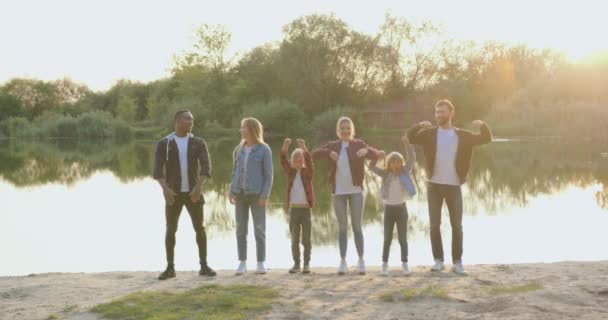 Primo piano di bonario sorridente multirazziale adulti e bambini che in piedi sulla riva del lago con le mani alzate e guardando la fotocamera con volti felici — Video Stock