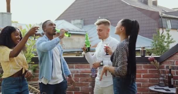 Atractiva sonriente feliz joven mixta gente divirtiéndose juntos durante la fiesta de amigos en la terraza con vistas a los tejados de la ciudad, cámara lenta — Vídeo de stock