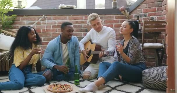 Портрет доброзичливої молодості безтурботної змішаної раси, яка розважається разом під час літньої вечірки друзів на балконі з піцою, напоями та піснями під гітарою — стокове відео