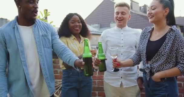 Gros plan de joyeux joyeux jeunes amis de course mixte insouciants accrochant la bière en bouteille et des verres avec du vin pendant la fête sur le balcon en été — Video