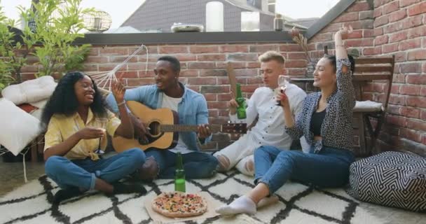 Portrait of likable positive happy young multiracial friends which singing songs at guitar,drinking beer and wine and having fun together during summer party on the terrace — Stock Video