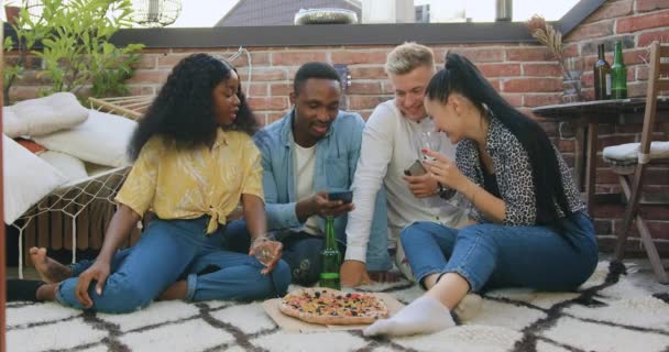 Vue de face de beau dans la bonne humeur moderne jeunes amis multiethniques qui sont assis sur le tapis dans le balcon et en utilisant leurs smartphones plus pendant la fête d'été — Video