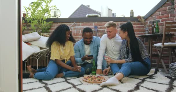Portrait de beaux amis multiethniques modernes souriants assis sur le tapis sur le balcon et utilisant le téléphone pendant la fête d'été — Video