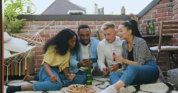 Vista frontal de amigos jóvenes multiétnicos guapos y de buen humor que se divierten juntos mientras revisan el videojuego en el teléfono inteligente y celebran la victoria durante la fiesta de verano en la terraza — Vídeos de Stock