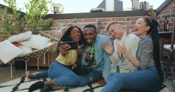 Ймовірно, щаслива весела багатоетнічна молодь розважається разом, а відеодзвінки по телефону зі спільним другом під час зустрічі друзів на терасі — стокове відео