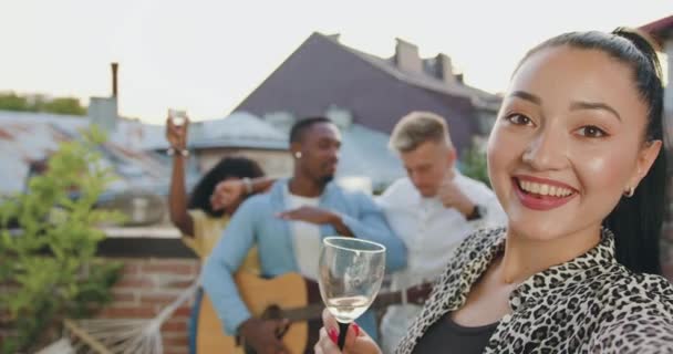 Hermosa mujer asiática joven sonriente con copa de vino haciendo selfie en el fondo de alegres amigos de raza mixta de buen aspecto en la terraza — Vídeos de Stock