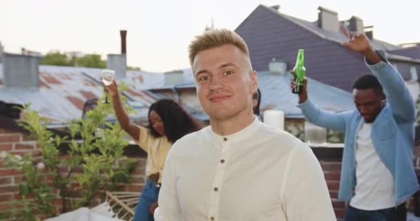 Knappe glimlachende jonge blonde man poseren op camera in de buurt van zijn positieve gemengde ras dansen vrienden op het terras tijdens de zomer feest, slow motion — Stockvideo