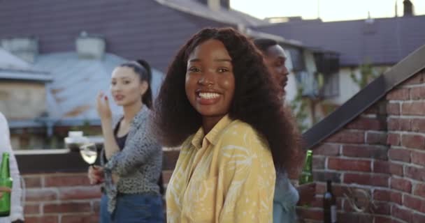 Вид спереду приваблива щаслива усміхнена молода чорношкіра дівчина, яка дивиться на камеру з задоволеним обличчям біля її позитивних безтурботних багаторасових друзів під час святкування літньої вечірки — стокове відео