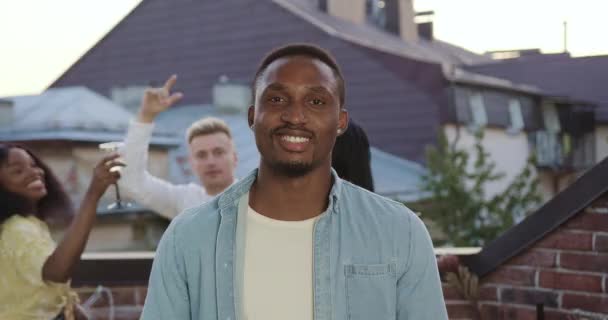 Güzel görünüşlü, gülümseyen Afrikalı Amerikalı yaz partisinde terasta dans eden arkadaşlarının yanında kameraya bakıyor. — Stok video