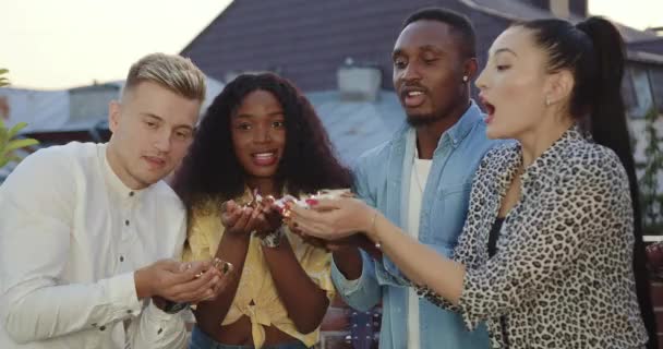 Front view of positive satisfied mixed race friends which blowing brilliant confetti during celebrating friends meeting on terrace at sundawn — Stock Video