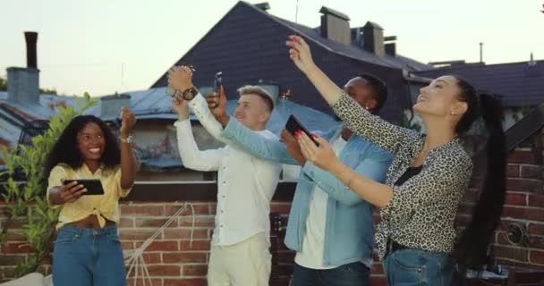 Jovens amigos de raça mista modernos e bem parecidos comemorando a festa de verão com brilhos no terraço com vista para os telhados da cidade ao pôr do sol — Vídeo de Stock