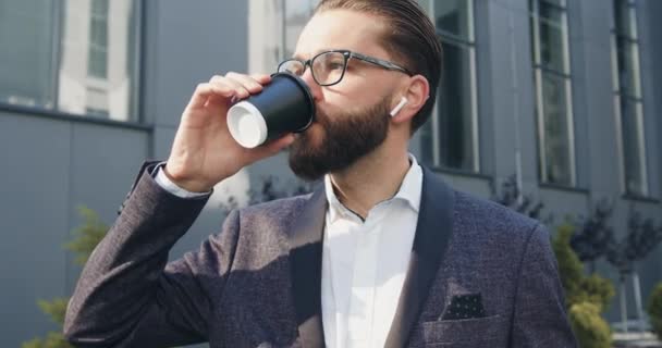 Primo piano di bell'aspetto elegante uomo barbuto pensieroso in bicchieri che bevono caffè vicino all'edificio urbano moderno mentre va al suo lavoro, 4k — Video Stock