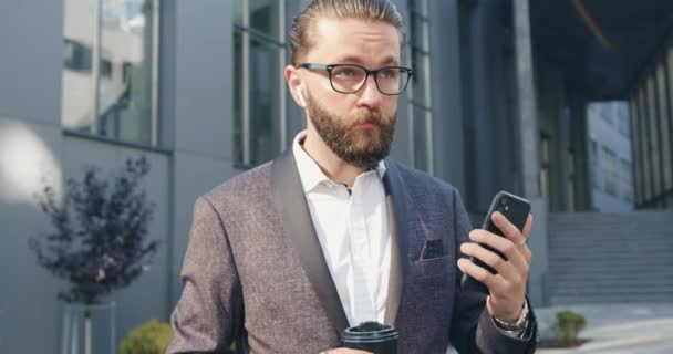 Guapo exitoso gerente de oficina barbudo adulto serio en gafas revisando sus airpods antes de comenzar a hablar en el teléfono inteligente cerca del moderno edificio de oficinas — Vídeos de Stock