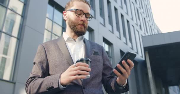 オフィスビルの近くにある電話プレイリストから、コーヒーを飲みながらエアポッドで音楽を聴くビジネススーツの35歳のスタイリッシュな髭の男の肖像 — ストック動画