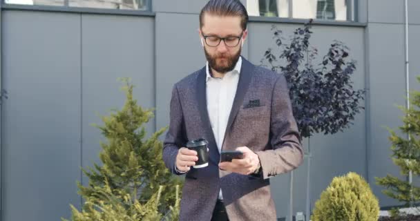 Vista frontale di attraente moderno uomo barbuto fiducioso in abito elegante che si distinguono all'aperto e parlando al telefono utilizzando airpods — Video Stock