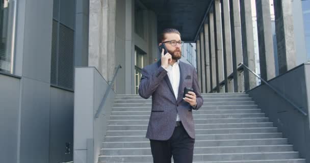 Gros plan d'attrayant sérieux confiant 35 ans directeur de bureau barbu avec des lunettes dans les vêtements de travail qui boire du café et parler sur smartphone tout en sortant de l'immeuble de bureaux — Video