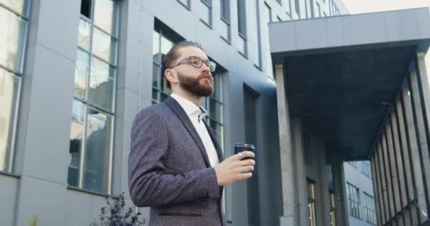 Guapo hombre de negocios barbudo preocupado de 35 años bebiendo café cerca del edificio de oficinas y comprobando el tiempo mientras espera a su socio de negocios, cámara lenta — Vídeo de stock