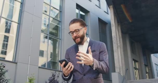 Вид спереду приємний у високому дусі успішний дорослий бородатий офісний працівник в окулярах, який здивований хорошими новинами на екрані телефону, стоячи біля сучасної будівлі — стокове відео