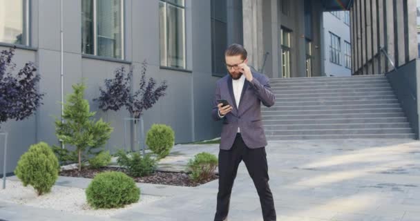 Vue de face de heureux heureux homme d'affaires confiant et confiant adulte barbu dans des lunettes et des vêtements formels qui obtenir de bonnes nouvelles sur son téléphone et se réjouir près de l'immeuble de bureaux — Video