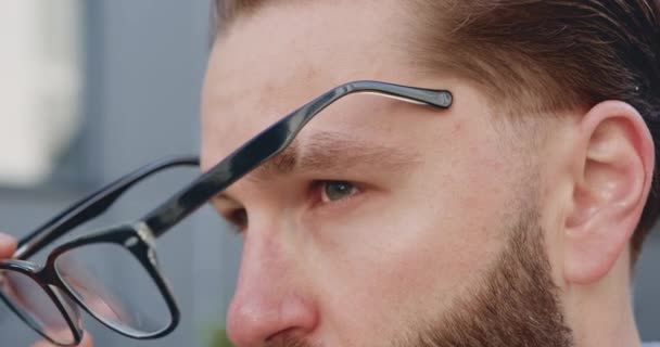 Porträt eines gutaussehenden ernsten selbstbewussten jungen bärtigen Mannes mit grünen Augen, der seine Brille aufsetzt und in die Ferne blickt — Stockvideo