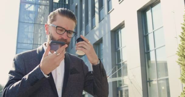 大規模な近代的なビジネスセンターの近くに歩いている間にスマートフォンで話して眼鏡や正式な服で成功した目的の髭のビジネスマンの可能性休憩時に — ストック動画