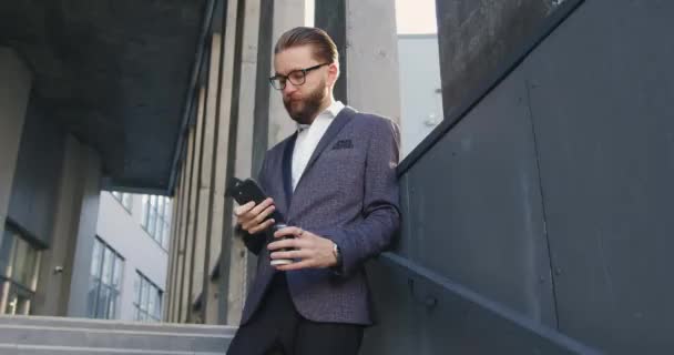 Крупный план симпатичного концентрированного успешного взрослого бородатого офисного менеджера в деловой одежде и очках, который использует свой мобильный телефон на ступеньках бизнес-центра — стоковое видео