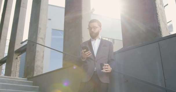 Charmant homme d'affaires équilibré de 35 ans, très qualifié, portant des lunettes et utilisant son smartphone pendant une pause café sur les marches d'un immeuble de bureaux — Video